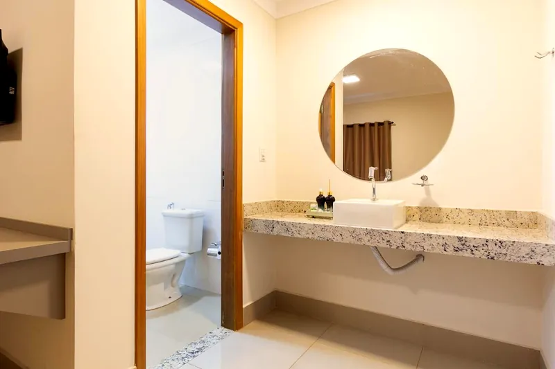 Banheiro do quarto com bancada de granito, cuba branca e espelho redondo