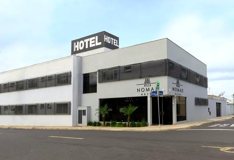 Fachada branca do Villa Nomad Hotel, com o letreiro HOTEL no alto, o logotipo Villa Nomad na entrada e palmeiras na calçada