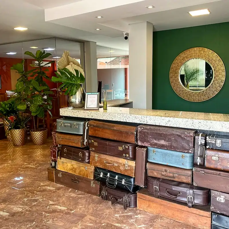 Recepção vista de lado: balcão de malas antigas, plantas em vasos dourados e parede verde com espelho