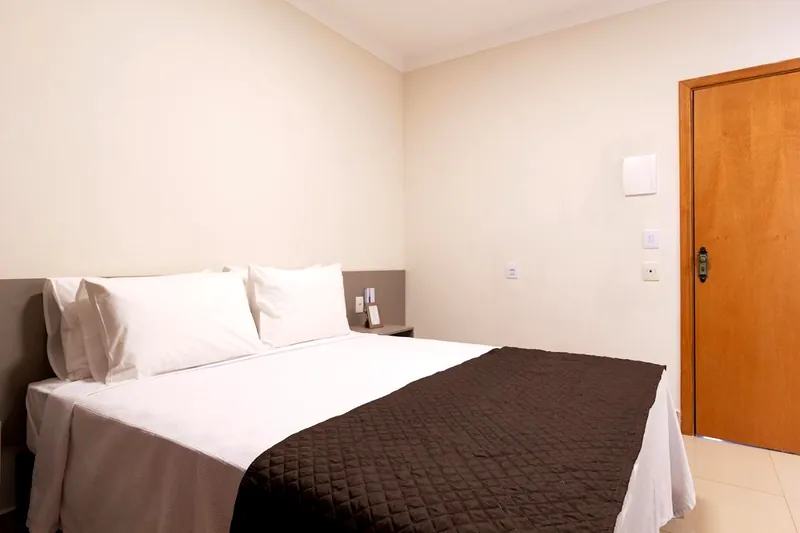 Cama de casal arrumada com travesseiros brancos e peseira marrom, ao lado da porta de madeira do quarto