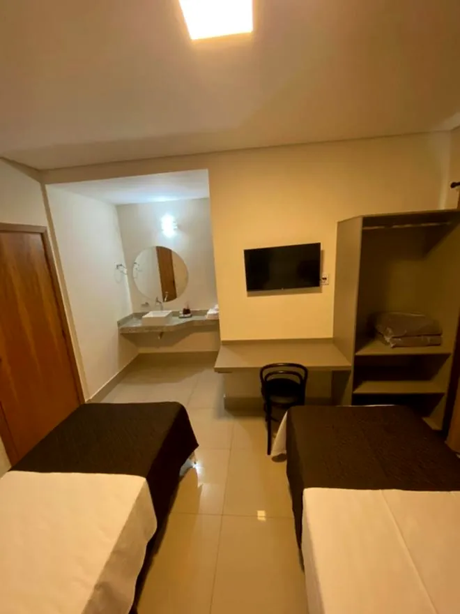 Vista do quarto a partir da porta, com camas, TV na parede, armário aberto e pia com espelho